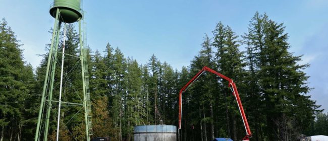 A photo of the new Quilcene concrete water tank still partial led in forms with the old standing metal water tank beside it. A concrete pump truck arm extends over the concrete tank for the final layer of forming.