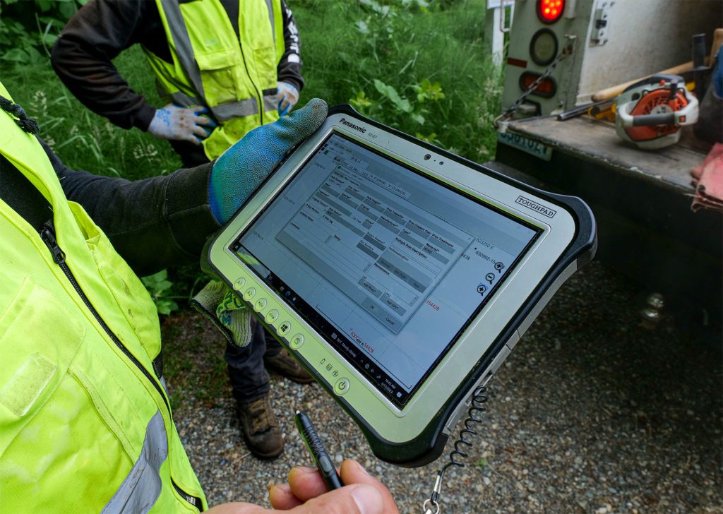 Worker in yellow vest holds a tablet and completes an inspection for a pole near the PPI truck.